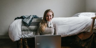 white woman sitting on the floor beside a bed with a laptop